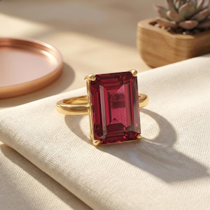 Pode incluir: Um anel de ouro com uma grande pedra preciosa retangular, vermelho escuro. O anel está sobre um tecido de cor neutra, com uma bandeja cor de cobre e uma pequena planta suculenta no fundo.