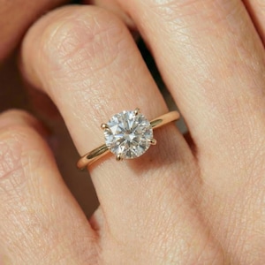 May include: A gold ring featuring a single round brilliant-cut clear gemstone set in a four-prong setting. The band is smooth and polished, showcasing a classic solitaire design with a high-shine metallic finish and a faceted center stone.