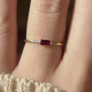May include: A thin gold ring featuring a rectangular baguette-cut deep red gemstone set horizontally. To the left of the central red stone, the band is accented with three tiny, clear, round-cut stones embedded in the metal shank.