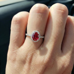 May include: A silver-toned ring featuring a central oval-cut vibrant red gemstone surrounded by a halo of small, clear, sparkling stones. The band is thin and also encrusted with tiny clear stones, creating a bright, reflective appearance.