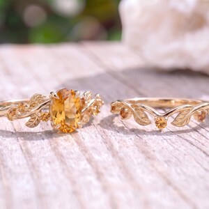 May include: A gold ring set featuring an oval-cut yellow citrine center stone flanked by engraved leaf motifs and small round accent stones, paired with a matching V-shaped gold band adorned with delicate leaves and tiny round citrine stones.