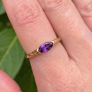 Könnte beinhalten: Ein goldener Ring mit einem einzelnen, marquise-geschliffenen violetten Amethyst-Edelstein, der in ein poliertes Metallband eingefasst ist. Der Stein ist horizontal in einer sicheren Fassung platziert und zeigt einen tiefvioletten Farbton.