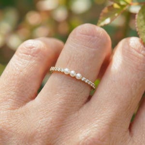 Op de afbeelding: Een gouden ring met drie kleine parels in het midden, omgeven door een band van kleine, heldere edelstenen. De ring zit om een vinger, met een vage groene en bruine achtergrond.