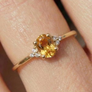 May include: A gold ring featuring a central oval-cut yellow citrine gemstone. The citrine is flanked on both sides by a cluster of three small, clear, round-cut white diamonds set into the gold band, creating a delicate and refined appearance.