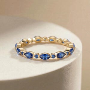 May include: A gold ring with a continuous band of alternating marquise-cut and round blue gemstones. The ring is set against a neutral background, highlighting the vibrant blue stones and the warm gold metal.