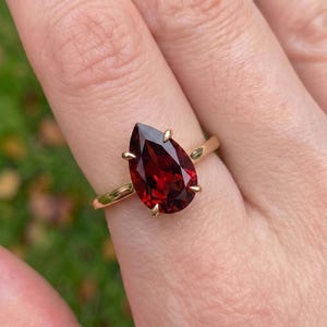 May include: A gold ring featuring a large, pear-shaped, deep red garnet gemstone held in place by four delicate prongs. The band is smooth and polished, showcasing a simple design that highlights the faceted stone's rich, dark crimson color.