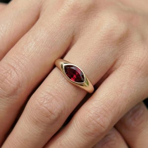 May include: A gold ring featuring a marquise-cut, deep red gemstone. The ring has a smooth, polished band and a bezel setting that highlights the vibrant color of the stone. The ring is shown on a person's finger.