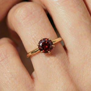 May include: A gold ring with a large, round, deep red gemstone. The gemstone is set with six prongs, and the band is a simple, polished gold. The ring is shown on a finger.