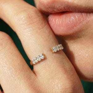 Puede incluir: Un anillo abierto de oro con dos segmentos distintos, cada uno engastado con una hilera de cuatro pequeñas piedras redondas y transparentes. La banda presenta un espacio entre las dos secciones con piedras, creando un diseño moderno.