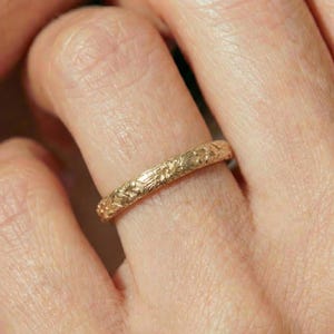May include: A gold ring with a floral pattern. The ring is worn on a finger, showcasing the intricate details of the embossed design. The ring's warm gold color contrasts with the skin tone, highlighting the delicate floral pattern.