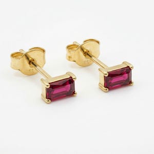May include: A pair of stud earrings featuring rectangular baguette-cut vibrant pink gemstones set in polished yellow gold. Each earring has a secure post back mechanism, showcasing a clean, metallic finish and a bright, saturated colour profile.