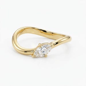 May include: A gold ring with a marquise-cut diamond. The ring's band curves in a flowing design, with the diamond set at the front. The ring is set against a white background, highlighting its polished finish.