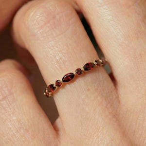 May include: A gold ring with alternating oval and round red gemstones. The ring's band is adorned with a repeating pattern of small, vivid red stones. The ring is worn on a finger, showcasing the intricate design and colour.