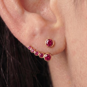 May include: A gold ear climber earring featuring six round, bezel-set vibrant red rubies that follow the curve of the earlobe. The design includes a single stud at the top and a graduated row of five smaller rubies extending along the lower edge.