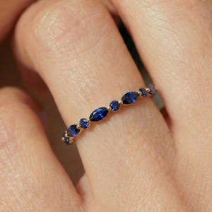 Op de afbeelding: Een gouden ring met afwisselend ronde en marquise-geslepen blauwe edelstenen. De ring wordt aan een vinger gedragen en toont het delicate ontwerp en de levendige kleur van de edelstenen.