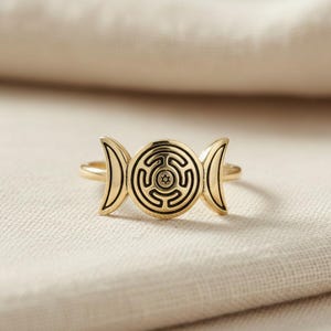 May include: A gold-toned ring featuring a central circular motif with an engraved labyrinth pattern and a small star at its core, flanked by two crescent moon shapes on either side. The metal surface is polished and smooth with dark engraved lines.