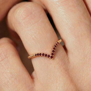May include: A V-shaped ring crafted from rose gold, featuring a series of small, round, deep red rubies set along the top edge of the band. The ring follows a chevron silhouette, with the gemstones arranged in a continuous line.