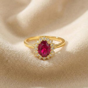 May include: A gold ring featuring a central oval-cut deep red gemstone surrounded by a halo of small, clear, round-cut stones. The band is smooth and polished gold, creating a classic cluster design that highlights the vibrant red center stone.