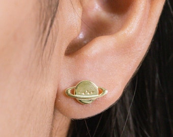 Puede incluir: Pendientes de color dorado en forma de planeta con un anillo alrededor. Los pendientes son pequeños y delicados.