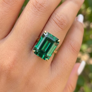 May include: A gold ring with a large, emerald-cut green gemstone. The ring has a simple gold band with four prongs holding the rectangular stone. The gemstone is a deep green colour, with visible facets.