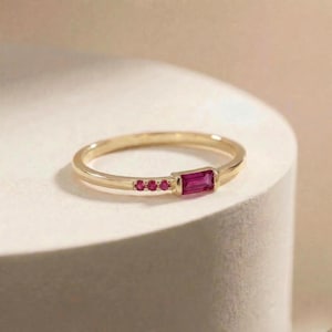 May include: A gold ring featuring a single rectangular baguette-cut pink gemstone and three small round pink gemstones set in a row along the thin band. The metal has a polished finish, and the stones are securely held in place by metal settings.