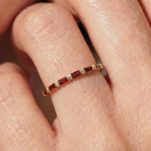 May include: A thin gold band ring featuring a series of small, rectangular, deep red baguette-cut gemstones set horizontally in a row. The metal setting between each stone is textured with fine vertical lines, creating a repeating pattern.