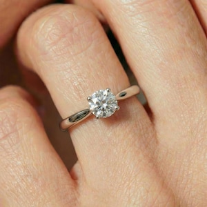 Puede incluir: Un anillo de metal plateado con una gema transparente de talla redonda engastada en cuatro garras. La banda es lisa y pulida, mostrando un diseño solitario clásico que rodea el dedo con un acabado metálico y reflectante.