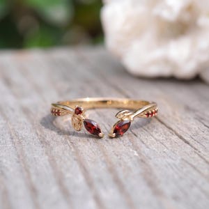 May include: A gold open ring featuring a leaf-inspired design with marquise-cut and round-cut red garnet gemstones. The band is crafted with delicate metal foliage detailing, showcasing two prominent marquise-cut garnets at the open ends.