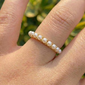 May include: A gold-colored ring featuring a continuous row of small, round, white pearls. The ring is worn on a finger, with a blurred green background.