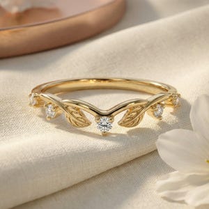 May include: A gold ring featuring a V-shaped band adorned with small, engraved leaf motifs and five round, clear, faceted stones set along the top edge. The metal has a polished finish, and the design follows a symmetrical, nature-inspired pattern.