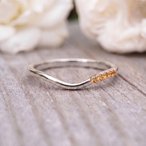 Puede incluir: Un anillo de banda plateada curvada con cinco pequeñas piedras preciosas de citrino redondas de color amarillo anaranjado engastadas a lo largo de un lado. El metal tiene un acabado pulido y reflectante, con una forma ondulada.