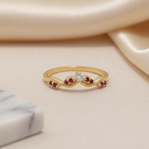 May include: A gold ring featuring a V-shaped band with a central clear round gemstone flanked by four small red gemstones on each side. The metal band has a smooth, polished finish and a curved design that tapers toward the center point.