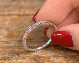 May include: A thin silver-toned metal ring featuring a continuous band of small, clear, sparkling stones set along the outer edge. The band has a textured, notched design along the sides, creating a repeating pattern that encircles the entire ring.
