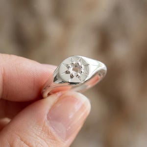 May include: A polished silver-tone signet ring featuring a circular face with an engraved eight-pointed star motif. The centre of the star contains a small, circular recessed setting designed to hold a single stone, surrounded by raised metal beads.