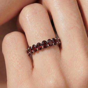 Puede incluir: Un anillo de plata con una serie de piedras preciosas ovaladas de color rojo intenso. Las piedras están engastadas muy juntas, creando una banda continua de color. El anillo se muestra en un dedo, destacando el diseño y el vibrante tono rojo de las piedras.