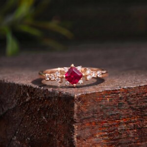 Può includere: Un anello in oro rosa con un rubino rosso intenso a taglio quadrato al centro. La fascia è impreziosita da piccoli diamanti trasparenti a taglio rotondo disposti in grappoli su entrambi i lati della pietra centrale, creando un design simmetrico.