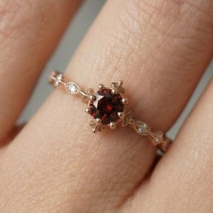 May include: A rose gold ring featuring a round, deep red gemstone at the center, held by a prong setting with decorative metal accents. The band is thin and delicate, adorned with small, clear, round stones set in a repeating pattern.
