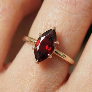 Puede incluir: Un anillo de oro con una gema roja oscura con corte marquesa. La gema está sujeta por cuatro garras. La banda del anillo es de oro pulido. El anillo se muestra en un dedo.
