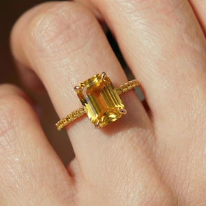 May include: A ring featuring a large, rectangular emerald-cut yellow gemstone held by four prongs. The band is crafted from a metallic material and is adorned with a series of small, round yellow stones set along the shank on both sides.