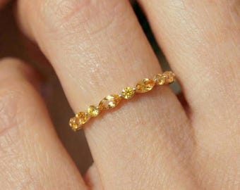 May include: A gold band ring featuring a series of alternating marquise-cut and round-cut yellow gemstones set in a continuous row. The ring has a polished metal finish and showcases a delicate, symmetrical arrangement of faceted stones.