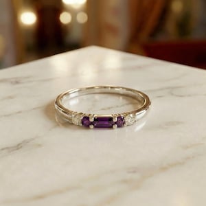 Op de afbeelding: Een zilverkleurige ring met een centrale rechthoekige paarse edelsteen in baguette-slijpvorm, geflankeerd door twee kleinere ronde paarse edelstenen en twee ronde heldere stenen, gezet in een gepolijste metalen band met een glad profiel.