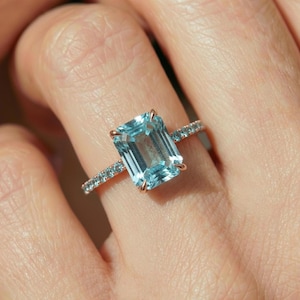 May include: A rose gold ring featuring a large, rectangular, light blue gemstone as the centerpiece. The band is adorned with smaller, matching blue stones, creating a sparkling effect. The ring's design is elegant and eye-catching.