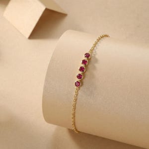 May include: A delicate gold chain bracelet featuring five round, bezel-set red rubies arranged in a vertical row. The bracelet is crafted with a fine link chain and polished gold metal settings that secure each vibrant gemstone in place.