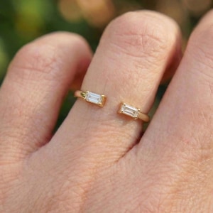 May include: A gold-colored open ring featuring two rectangular, clear gemstones. The ring is set on a finger, showcasing the minimalist design and the sparkle of the stones. The ring's design is modern and elegant.