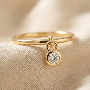 May include: A gold ring featuring a thin, smooth band and a single dangling charm. The charm consists of a small, circular gold frame holding a clear, faceted round gemstone that catches the light, suspended from a small gold loop on the band.