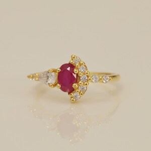 May include: A gold ring featuring a central oval-cut deep red ruby gemstone. The ruby is flanked by a pear-cut clear stone on one side and a curved arrangement of small, round clear stones on the other, all set on a thin, polished gold band.