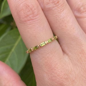 Può includere: Un sottile anello a fascia in oro caratterizzato da una serie di piccole pietre preziose rettangolari verde chiaro con taglio baguette, disposte orizzontalmente e separate da piccole barre in metallo dorato in un motivo ripetitivo.