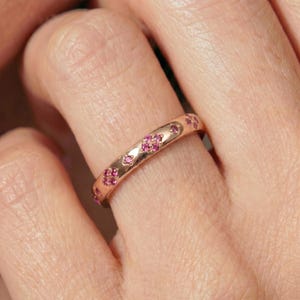 May include: A rose gold band ring featuring a series of small, circular, pink gemstone clusters set at intervals along the outer surface. The metal has a polished finish, and the stones are arranged in groups of four to create a floral-like pattern.