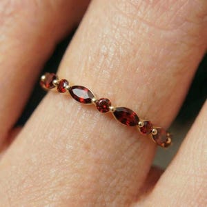 May include: A gold ring featuring alternating oval and round red gemstones. The ring's band is adorned with a series of gemstones, forming a continuous pattern. The ring is displayed on a finger.