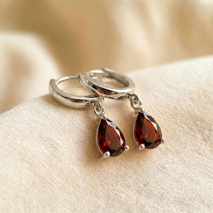 May include: A pair of silver-toned hoop earrings featuring dangling, pear-shaped, deep red garnet gemstones. Each earring consists of a polished metal hoop with a faceted, teardrop-shaped stone suspended from a small jump ring at the base.
